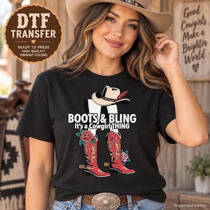 May include: A black t-shirt featuring a graphic print of a white cowboy hat, red cowboy boots with gold detailing, and pink roses. The text reads: BOOTS & BLING It's a Cowgirl THING. The design is centered on the chest of the short-sleeved garment.