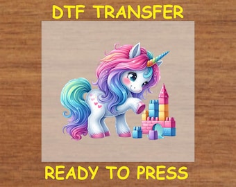 May include: A DTF transfer design featuring a colorful unicorn with a rainbow mane and tail, holding a toy building block. The unicorn is standing in front of a colorful toy castle made of blocks. The design is ready to be pressed onto a fabric item.