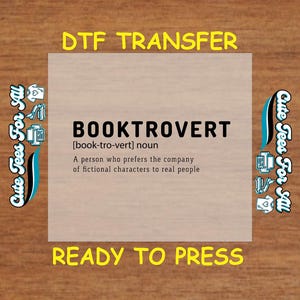 May include: A light brown wood background with a white square in the center. The square has a blue and white design on the left and right sides. The text "DTF TRANSFER" is in yellow at the top of the square. The text "BOOKTROVERT" is in black at the top of the square. The text "[book-tro-vert] noun" is in black below the word "BOOKTROVERT". The text "A person who prefers the company of fictional characters to real people" is in black below the noun definition. The text "READY TO PRESS" is in yellow at the bottom of the square.