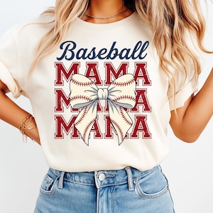 May include: Cream-colored t-shirt with the words "Baseball MAMA" in a stacked design. The word "Baseball" is in navy blue, and "MAMA" is in red. A baseball-themed bow graphic is centered on the shirt.