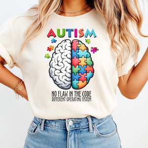 May include: Cream-colored t-shirt with the word "AUTISM" in colorful letters above a brain graphic. The brain is split, one side white, the other a puzzle. Text below reads "NO FLAW IN THE CODE DIFFERENT OPERATING SYSTEM."