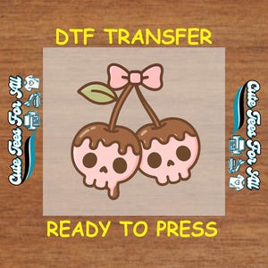 May include: DTF transfer featuring a pair of cartoon cherries with skull-shaped faces, topped with chocolate and a pink bow. The text "DTF TRANSFER" and "READY TO PRESS" are in yellow. The words "Cute Tees For All" are on the sides.
