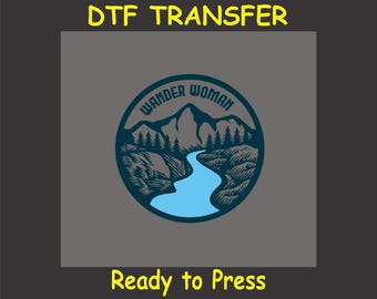 Könnte beinhalten: Ein blaues und weißes kreisförmiges Design mit dem Text "Wander Woman" und einer Bergszene mit einem Fluss, der durch sie fließt. Der Text "Ready to Press" befindet sich unter dem Design.