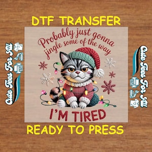 May include: A DTF transfer featuring a cartoon cat wearing a Christmas sweater and hat, adorned with string lights and snowflakes. Text includes "Probably just gonna jingle some of the way" and "I'M TIRED READY TO PRESS". The design is on a light brown background.