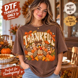 May include: A brown t-shirt featuring a graphic print of a turkey with a bow, surrounded by the word THANKFUL, pumpkins, acorns, and autumn leaves. The design includes patterns like leopard print, plaid, and floral motifs in orange and brown.