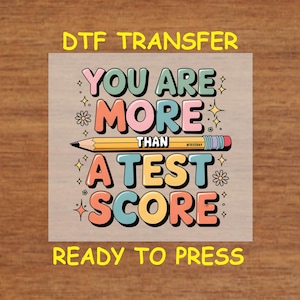 May include: DTF transfer with the text "YOU ARE MORE THAN A TEST SCORE" in colorful bubble letters, with a pencil graphic. The words "READY TO PRESS" are at the bottom. The background is a light tan color.