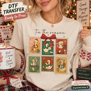 May include: A cream-colored t-shirt featuring a graphic print of six postage stamp-style illustrations of geese in Christmas scenes, with the text 'Tis the season' above and a large red bow centered at the top of the grid of stamps.