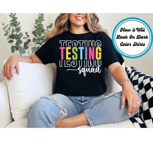 May include: Black t-shirt with the text "Testing Testing Testing Squad" in rainbow colors. The text is stacked vertically with each word in a different color.