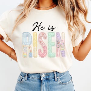 May include: Cream-colored t-shirt with the text "He is Risen" in black script. The word "Risen" is composed of pastel-colored patchwork letters, each with a unique pattern and floral accents, creating a soft, Easter-themed design.