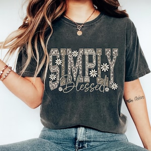 May include: Dark gray t-shirt with the words "SIMPLY Blessed" in a combination of leopard print and script font. White daisy accents are placed around the text. The shirt is a casual style.