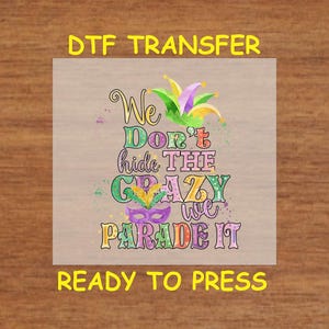 May include: DTF transfer with the text "We Don't Hide The Crazy We Parade It" in colorful letters. The design includes a jester hat, a mask, and confetti. The words "Ready to Press" are also included.