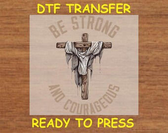 May include: DTF transfer graphic featuring a wooden cross with a draped white cloth. The text "BE STRONG AND COURAGEOUS" and "READY TO PRESS" are in a light brown color.