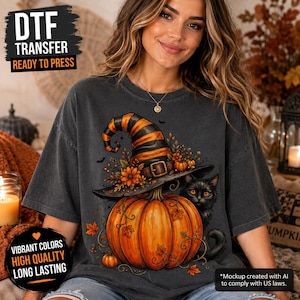 May include: A charcoal gray t-shirt featuring a graphic print of a large orange pumpkin topped with a black and orange striped witch hat, decorated with autumn flowers and leaves, accompanied by a black cat with bright orange eyes.
