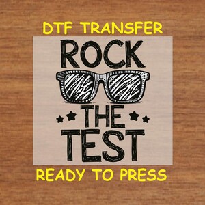 May include: Black and white graphic design with the text "Rock the Test" and a pair of sunglasses. The text "DTG Transfer Ready to Press" is below the graphic.