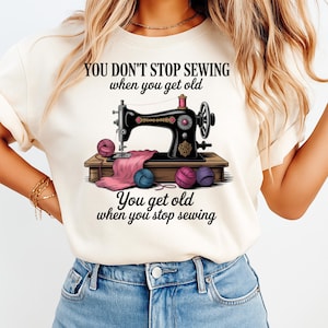 May include: Cream-colored t-shirt featuring a vintage sewing machine illustration. The black machine is on a wooden table with pink fabric and colorful yarn balls. Text reads "YOU DON'T STOP SEWING when you get old. You get old when you stop sewing."