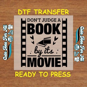 May include: A black and white design on a clear sheet, ready to be pressed onto fabric. The design says "Don't judge a book by its movie". The design is framed by a film strip.