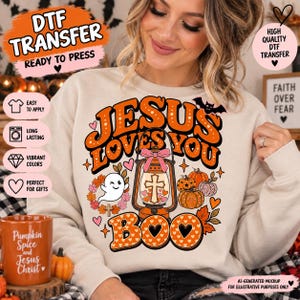 May include: A beige crewneck sweatshirt featuring a graphic print with the text Jesus Loves You Boo. The design includes an orange lantern with a cross, a pink bow, a winking ghost, pumpkins, autumn leaves, and a bat in orange and black.