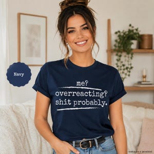 May include: A navy blue short-sleeved t-shirt featuring white text that reads: me? overreacting? shit probably. The shirt has a crew neckline and is worn with a black belt and blue denim jeans. A navy blue circular icon is visible on the left side.