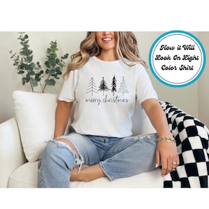 Puede incluir: Camiseta blanca con un dibujo lineal negro de cuatro &aacute;rboles de Navidad y la frase "merry christmas" en fuente manuscrita. La camiseta es de color claro y tiene mangas cortas.