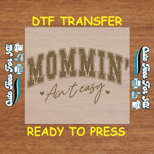 May include: A brown DTF transfer sheet with the text "MOMMIN' Ain't easy" in a distressed font. The sheet is ready to be pressed onto a garment.