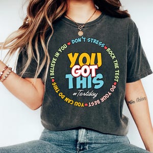 May include: Dark gray t-shirt with the text "YOU GOT THIS" in yellow, white, and blue. The phrase is surrounded by a circular design with the phrases "I BELIEVE IN YOU" and "DON'T STRESS ROCK THE TEST". The hashtag #Testday is also included.