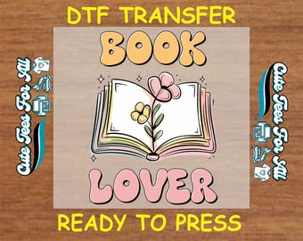 May include: A DTF transfer design featuring a book with a flower on top. The text "BOOK LOVER" is in pink and yellow bubble letters. The text "READY TO PRESS" is in yellow letters.