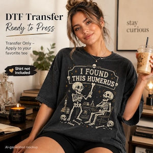 May include: A charcoal gray t-shirt featuring a graphic print of two skeletons at a desk with the text I FOUND THIS HUMERUS. The design includes small skulls, stars, and books. Text on the image reads DTF Transfer Ready to Press, Transfer Only - Apply to your favorite tee, and Shirt not included.