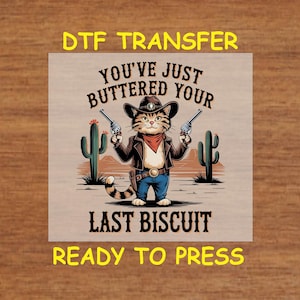 May include: A digital transfer design featuring a cartoon cat wearing a cowboy hat and holding two pistols. The text reads "You've just buttered your last biscuit" and "Ready to press".