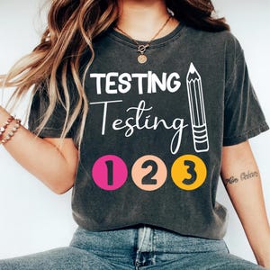 May include: Dark gray t-shirt with the words "TESTING Testing Testing" in white, along with a pencil graphic and the numbers 1, 2, and 3 in pink, peach, and yellow circles. The shirt is a casual style.