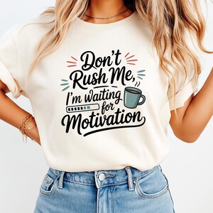 May include: Cream-colored t-shirt with the text "Don't Rush Me, I'm waiting for Motivation" in black script. The design includes a coffee cup and a loading bar graphic. The shirt is a casual wear item.