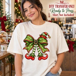 May include: A white t-shirt featuring a graphic of a green polka-dot bow with a gold buckle, attached to striped green and white elf legs wearing red curled shoes and a matching elf hat with a gold bell. Text reads DTF TRANSFER ONLY.