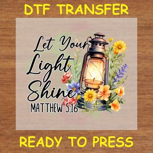 May include: A white iron lantern with a flame inside, surrounded by yellow and orange flowers. The text "Let Your Light Shine Matthew 5:16" is written in black cursive lettering.