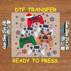 May include: A DTF transfer design featuring six colorful game controllers with holiday decorations. The controllers are red, white, green, and tan, with snowflakes, candy canes, and string lights. Text reads "DTF TRANSFER" and "READY TO PRESS."