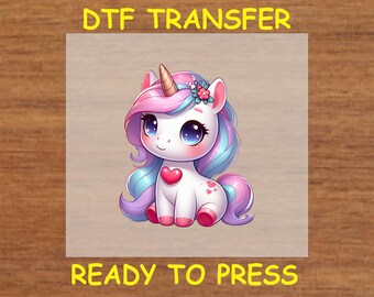 May include: A cute cartoon unicorn with a pink and blue mane and tail, wearing a pink flower crown. The unicorn has a pink heart on its chest and is sitting on a white background. The text "DTF TRANSFER READY TO PRESS" is visible at the top and bottom of the image.