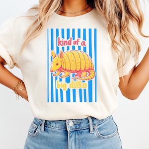 May include: A cream-colored short-sleeve t-shirt featuring a graphic of a yellow armadillo centered over vertical blue stripes. The text "kind of a" appears in pink above the armadillo, and "big dillo" appears in yellow below the graphic.
