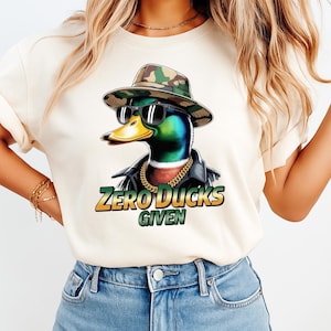 May include: Cream-colored t-shirt featuring a cartoon duck wearing sunglasses, a camouflage hat, and a gold chain. The duck is wearing a black leather jacket. The text "ZERO DUCKS GIVEN" is printed in green and gold.