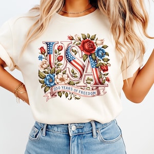 K&ouml;nnte beinhalten: Cremefarbenes T-Shirt mit dem Wort "USA" in einem patriotischen Design. Die Buchstaben integrieren die amerikanische Flagge und sind von roten, wei&szlig;en und blauen Blumen umgeben. Der Text "250 YEARS OF FREEDOM" befindet sich auf einem Banner.