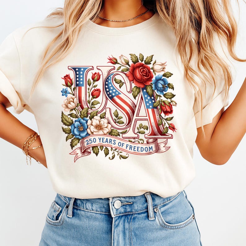 K&ouml;nnte beinhalten: Cremefarbenes T-Shirt mit dem Wort "USA" in einem patriotischen Design. Die Buchstaben integrieren die amerikanische Flagge und sind von roten, wei&szlig;en und blauen Blumen umgeben. Der Text "250 YEARS OF FREEDOM" befindet sich auf einem Banner.