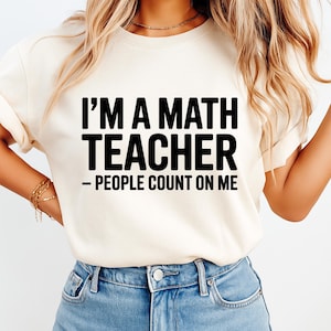 May include: A cream-colored short-sleeve t-shirt featuring the black text I'M A MATH TEACHER – PEOPLE COUNT ON ME printed on the front. The shirt has a crew neckline and is paired with blue denim jeans, creating a casual and straightforward appearance.