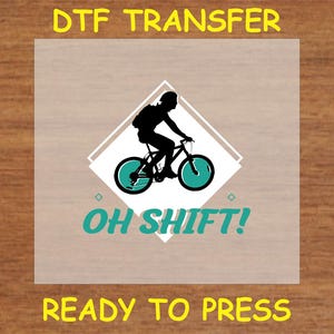 May include: A clear sheet with a black silhouette of a person riding a bicycle with teal wheels. The text "OH SHIFT!" is below the bicycle and the text "DTG TRANSFER READY TO PRESS" is at the top and bottom of the sheet.