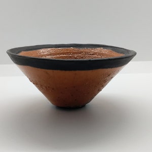May include: A small, terracotta-colored bowl with a black rim. The bowl is cone-shaped and has a slightly textured surface.