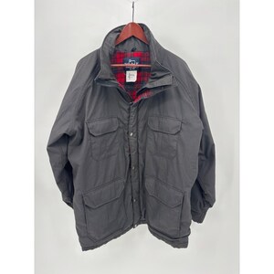 May include: A charcoal gray jacket featuring a high collar, a front zipper closure with snap buttons, and four large flap pockets on the front. The interior is lined with a red, black, and green plaid fabric, and a label reads Woolrich.