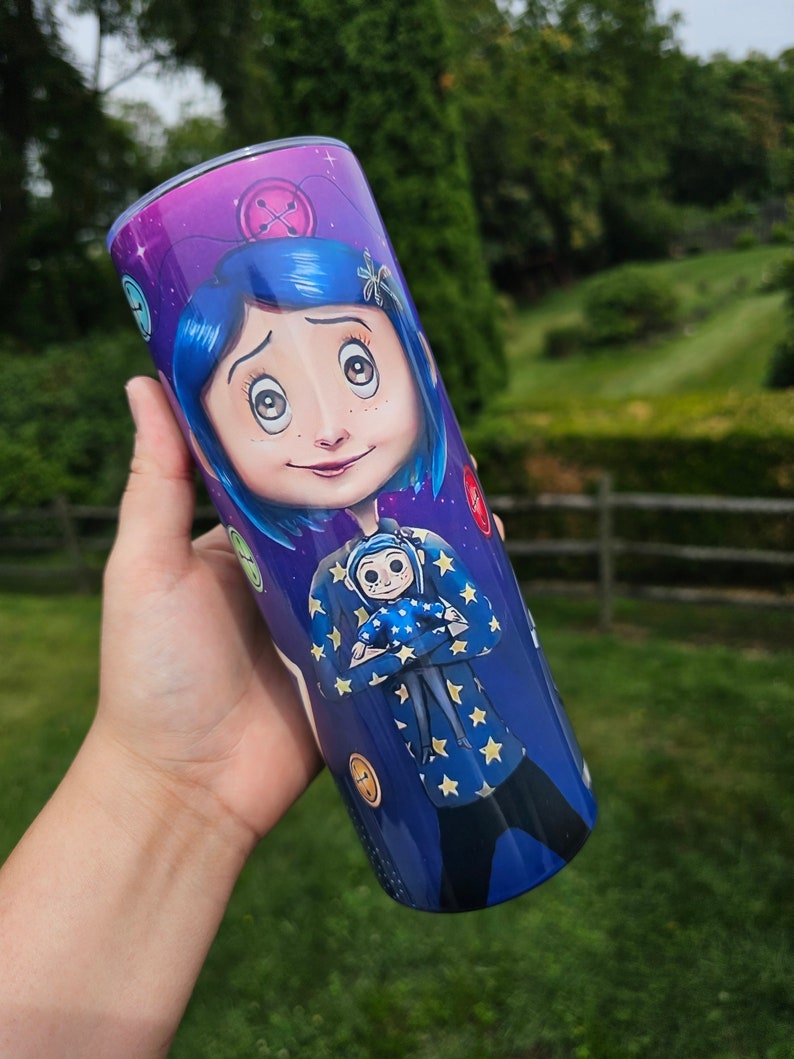 May include: A stainless steel tumbler featuring a vibrant illustration of a character with blue hair and a starry jacket. The design includes button and star motifs against a purple and blue background, creating a whimsical aesthetic.