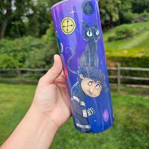 May include: A purple and blue tumbler featuring characters and button designs. The tumbler has a stainless steel rim and a clear lid. The design includes a black cat, a character in a blue jacket, and various button illustrations.