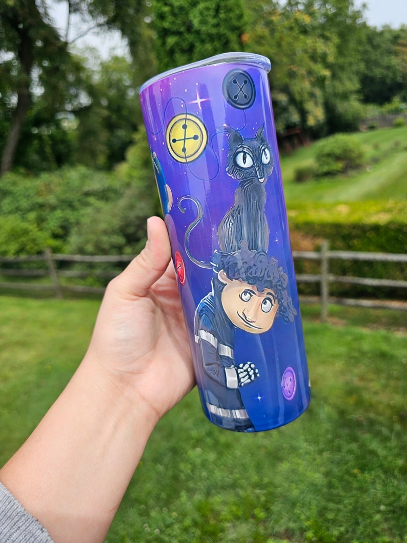 May include: A purple and blue tumbler featuring characters and button designs. The tumbler has a stainless steel rim and a clear lid. The design includes a black cat, a character in a blue jacket, and various button illustrations.