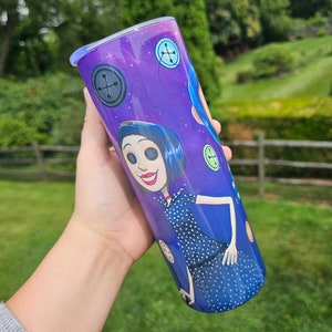 May include: A stainless steel tumbler featuring a cartoon character in a blue dress with white polka dots. The tumbler has a purple background with button and star graphics. The character has blue hair and is smiling. The tumbler is cylindrical and has a blue lid.