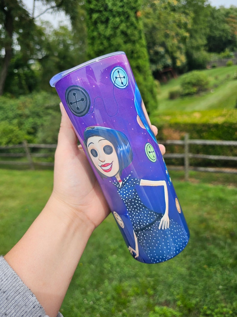 May include: A stainless steel tumbler featuring a cartoon character in a blue dress with white polka dots. The tumbler has a purple background with button and star graphics. The character has blue hair and is smiling. The tumbler is cylindrical and has a blue lid.