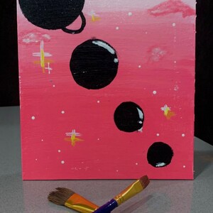 May include: A pink and white canvas painting with black circles and a ring around one of the circles. The painting is on a table with two paint brushes.