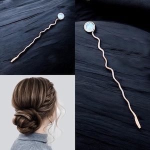 May include: A wavy, gold-toned hair stick with a clear, round crystal at the top. The stick is shown against a dark wood background and in a model's hair.