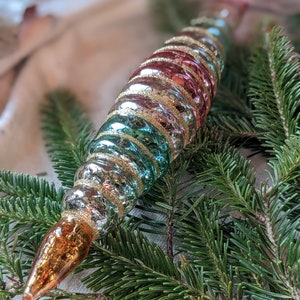 May include: A vintage glass Christmas ornament with a spiral design in shades of pink, teal, and gold. The ornament is decorated with glitter and has a pointed tip.